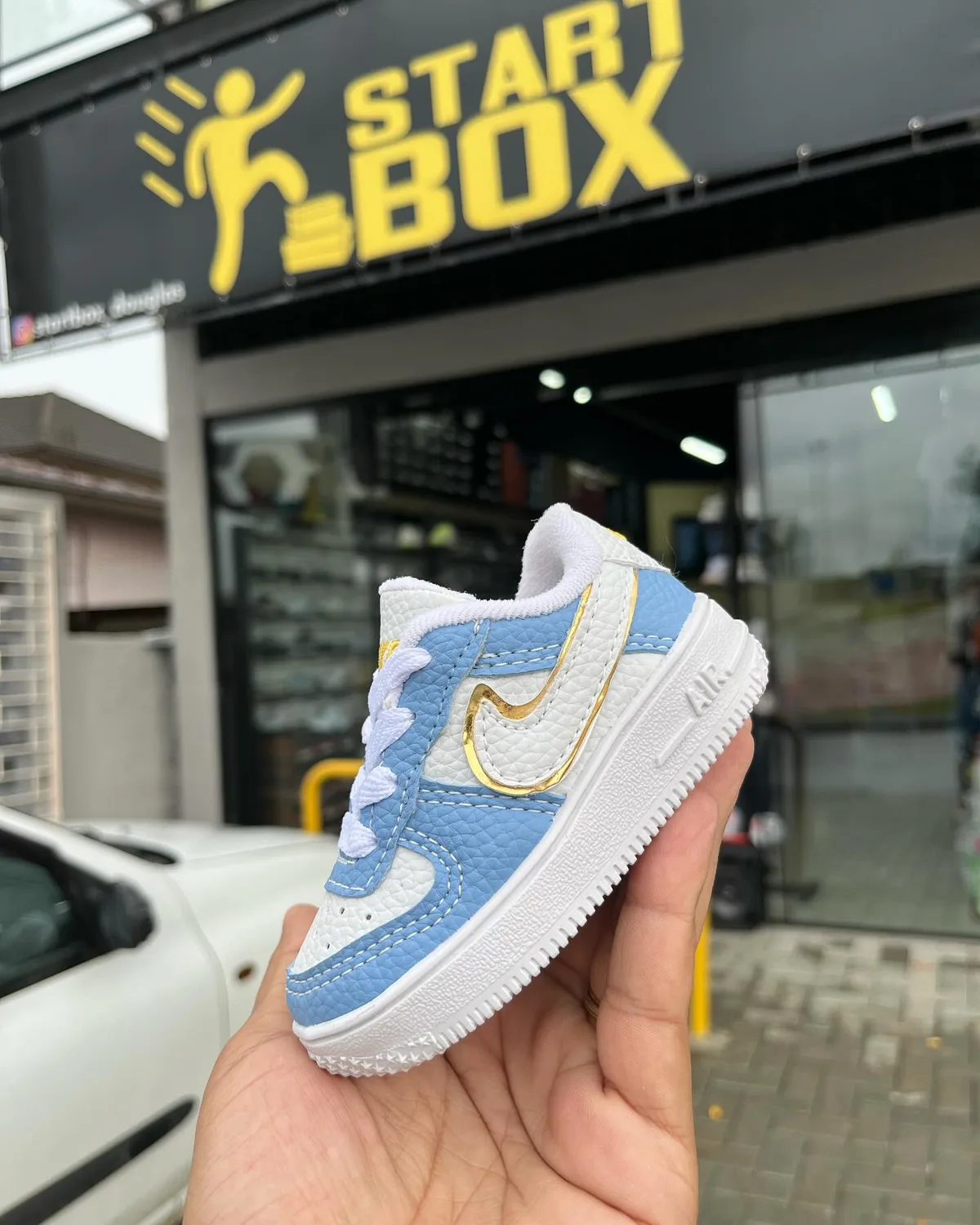 Um Air Force infantil branco e azul segurado na calçada, com a fachada da Startbox atrás: o letreiro preto com o START BOX amarelo e os frades amarelos